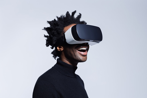 Portrait of excited afro american man wearing virtual reality glasses, standing against grey background.